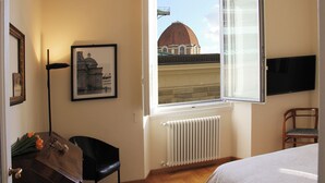 Una habitación con una ventana grande, un radiador, un cuadro enmarcado, una silla y una cama.