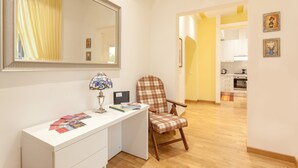 Ein Zimmer mit weißem Schreibtisch, einem Stuhl, einem Spiegel und Blick in einen Küchenbereich.