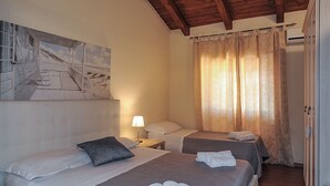 A hotel room with two beds, a painting of a beach scene, a wooden ceiling, and a window with curtains. - Katane Holidays "Verdi Apartment with Triple Room" (Catania)
