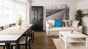 Una sala de estar moderna con comedor, que presenta un ventanal amplio, un cuadro de un puente y un sofá blanco con cojines de colores.