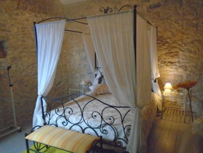 Rest in a traditional four-poster bed with a canopy, set against a stone wall, and enjoy a cozy reading area with a table and lamp.
