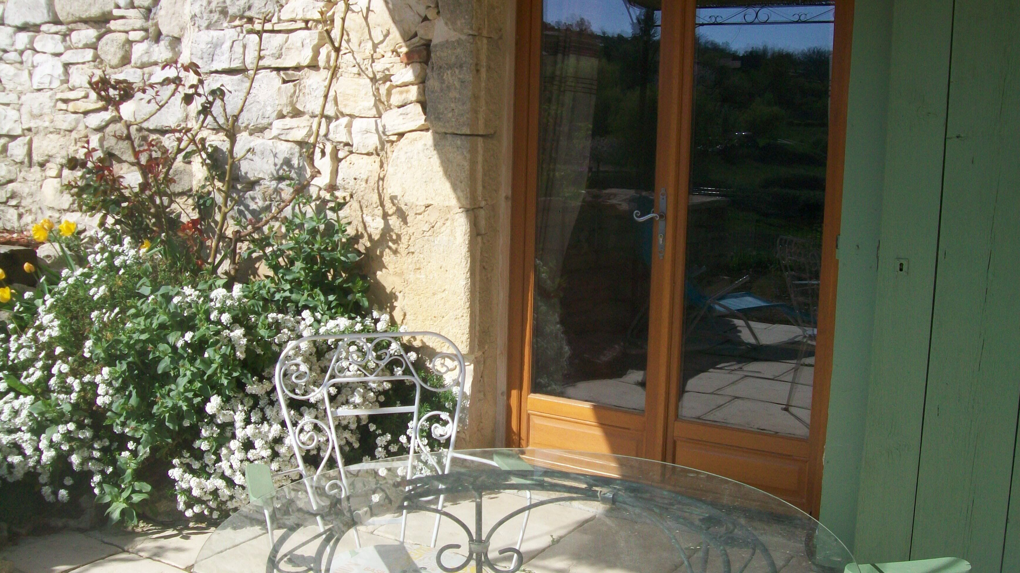 Taula de vidre amb cadires metàl·liques davant d'una paret de pedra amb plantes florejants.