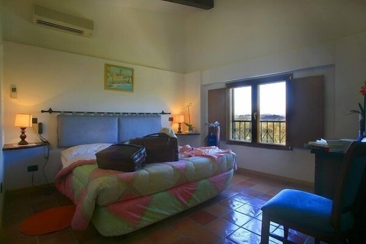 A bedroom with a bed, a chair, a window with a view, and a painting on the wall.