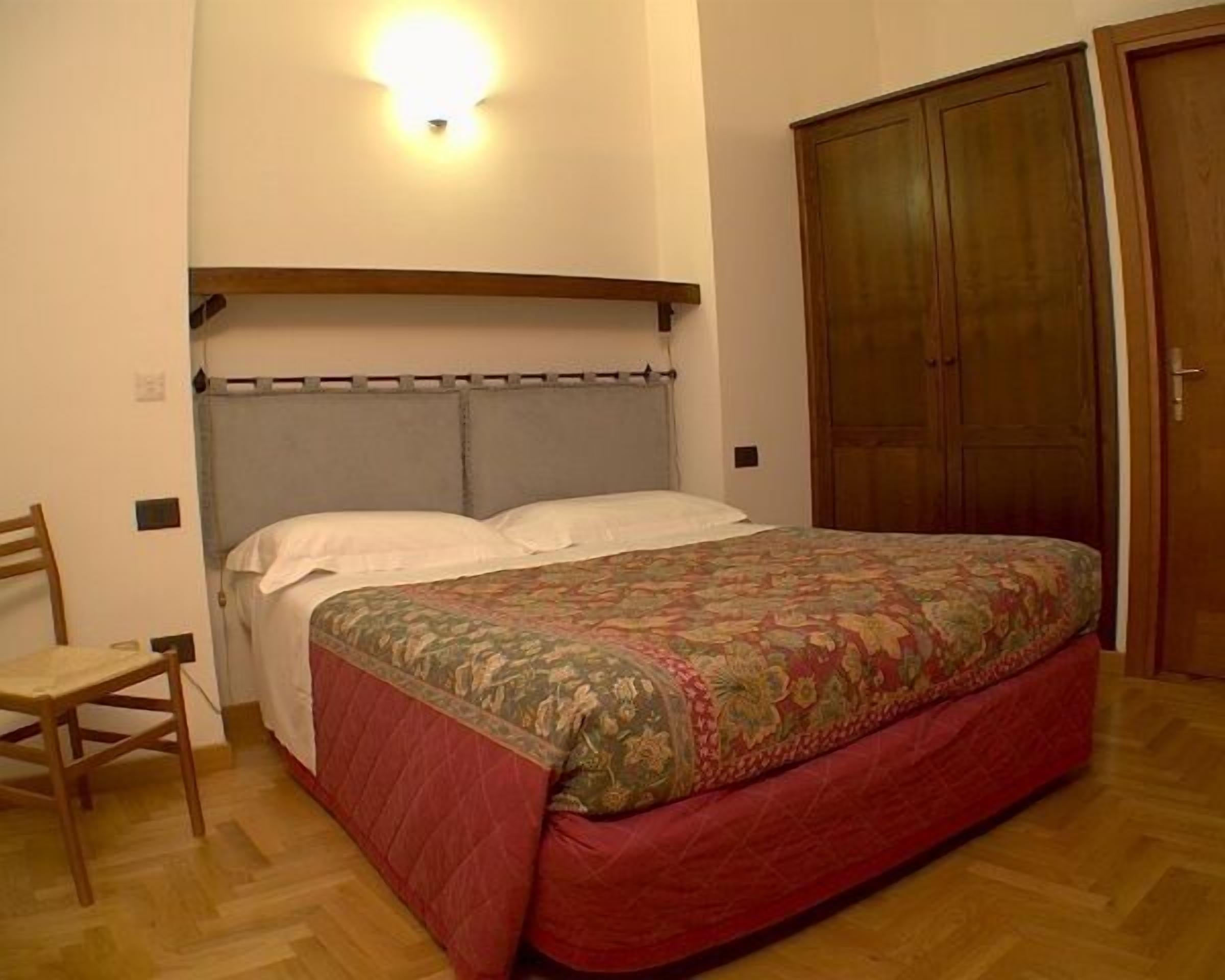 A bedroom with a bed, a wooden chair, a wardrobe, and a wall-mounted light.