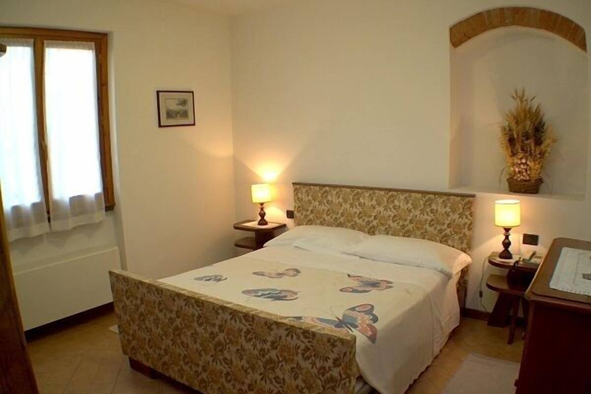 A bedroom with a bed, two bedside tables, a lamp on each table, a window with curtains, a wall-mounted picture, and a small desk with a lamp.