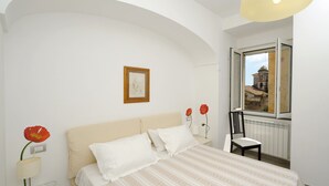 Une chambre à coucher comprenant un lit, une table de chevet, une chaise, une fenêtre donnant sur un immeuble et un tableau accroché au mur.