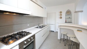 Une cuisine moderne équipée d’une cuisinière à gaz, d’un évier en acier inoxydable et de meubles blancs. On y trouve trois tabourets devant un comptoir-bar et une porte qui donne sur une autre pièce.