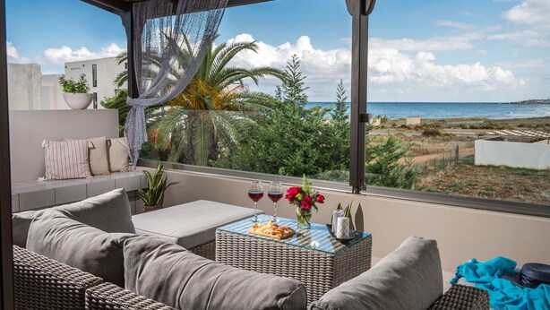 En balkon med fletmøbler, et bord med snacks og drikkevarer, og udsigt til hav og grønt.