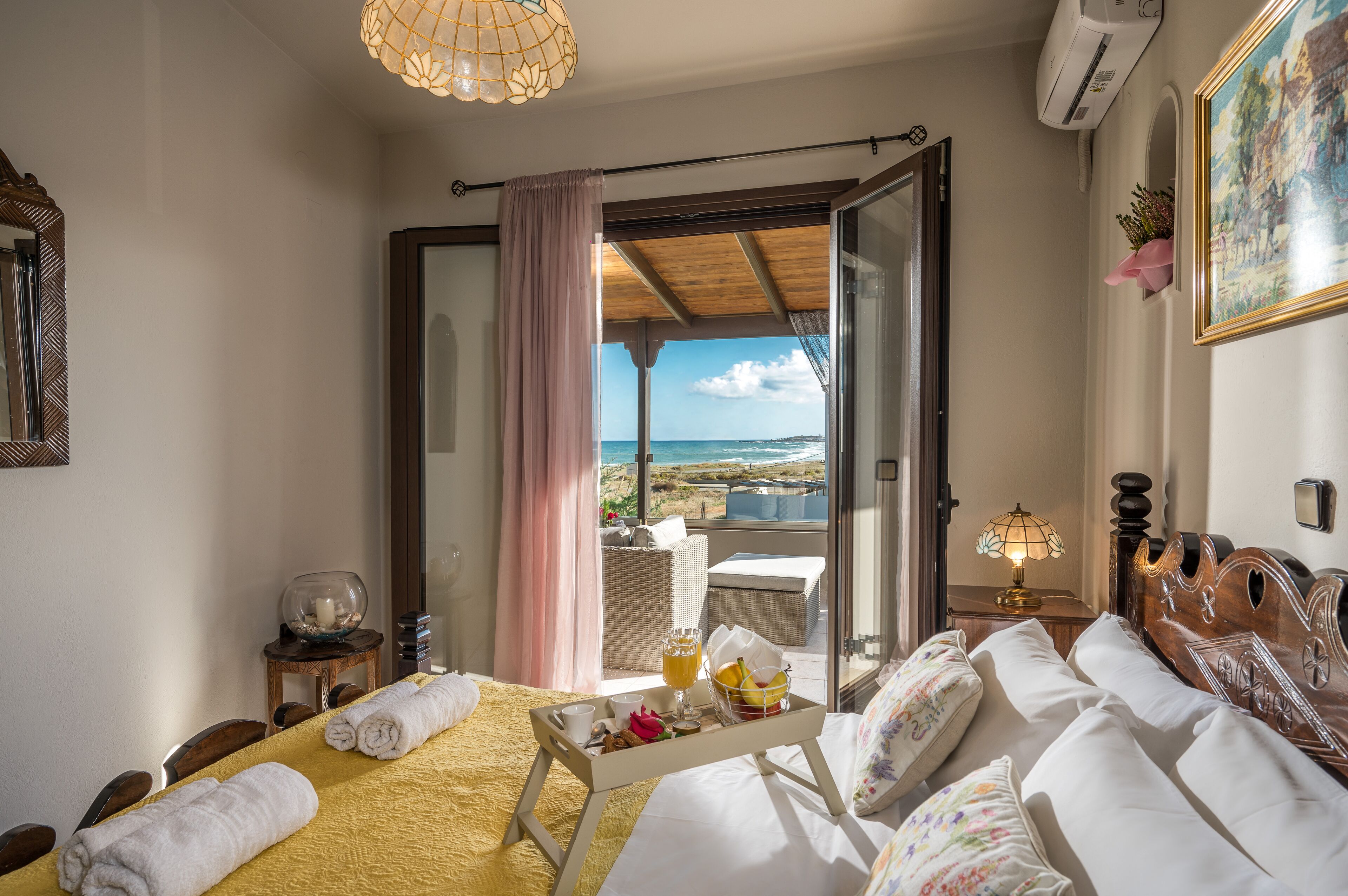 Habitación de hotel con cama, mesa con desayuno, vista a la playa y balcón con mobiliario de mimbre.