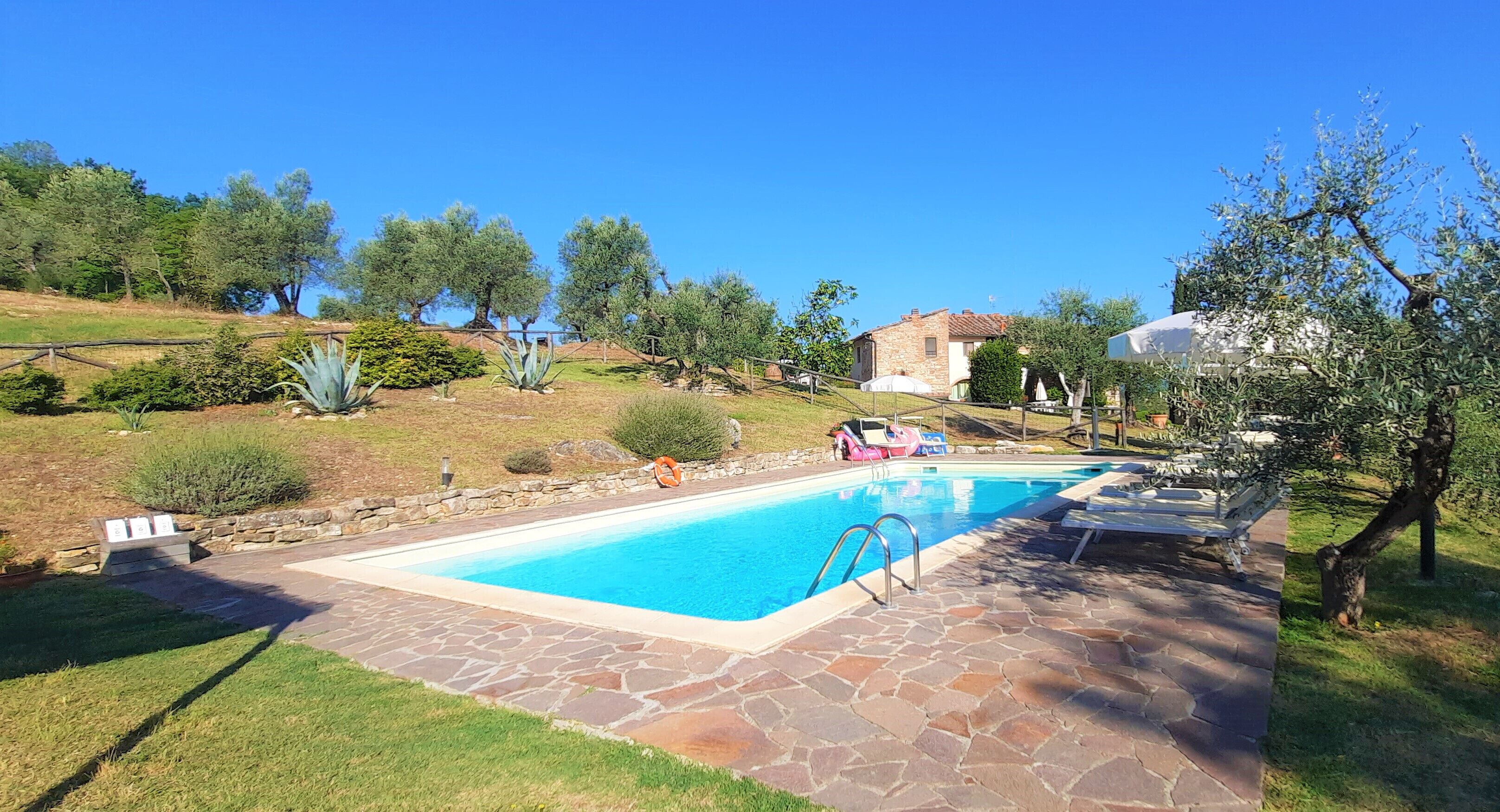 Una piscina con baldosas de piedra, rodeada de césped y árboles, con una casa de piedra al fondo.
