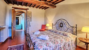 Une chambre avec un plafond en bois, un lit recouvert d’une literie à motifs floraux, une chaise rouge et une petite table avec une lampe.