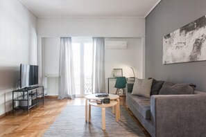 En moderne stue med en grå sofa, et fjernsyn på et skærmstativ, et sofabord og et stort vindue med gardiner.
