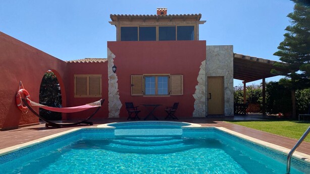 A swimming pool with a stone wall and a red building in the background. - Villa Nido Cari—Fuerteventura Rural Oasis (Casillas de Morales)