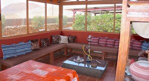 Relax in a serene outdoor space with a bench, cushioned seating, and a hookah, surrounded by lush greenery and distant hills.