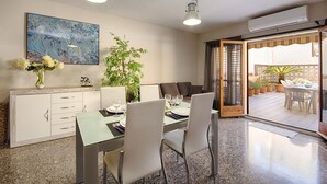 Een eetruimte met een tafel voor vier personen, een wit bergkast met lades, een groot schilderij aan de muur en een glazen schuifdeur die leidt naar een terras.