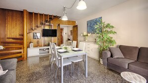 Een moderne woonkamer met een eethoek, houten lambrisering, een flatscreen televisie, een grijze bank en een potplant.