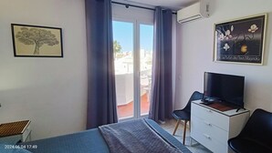 Une chambre avec un lit, un bureau, une chaise, une commode et un téléviseur. Il y a une fenêtre avec des rideaux, un tableau encadré au mur et un appareil de climatisation.