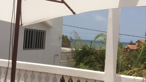 A balcony with a white railing, a window with vertical bars, and a view of the sea and buildings in the background. - Beachfront accommodation Yene - Senegal (Yene Kao)