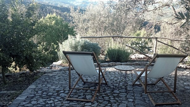 An outdoor seating area with two chairs and a small table, surrounded by a stone-paved patio and a view of a mountainous landscape. - Casa del Vento in San Cono, ancient stone dwelling among olive trees (Polizzi Generosa)