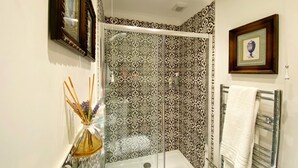 Une salle de bain avec une douche à parois de verre, des carreaux décorés, un tableau encadré et un vase en verre contenant des roseaux.