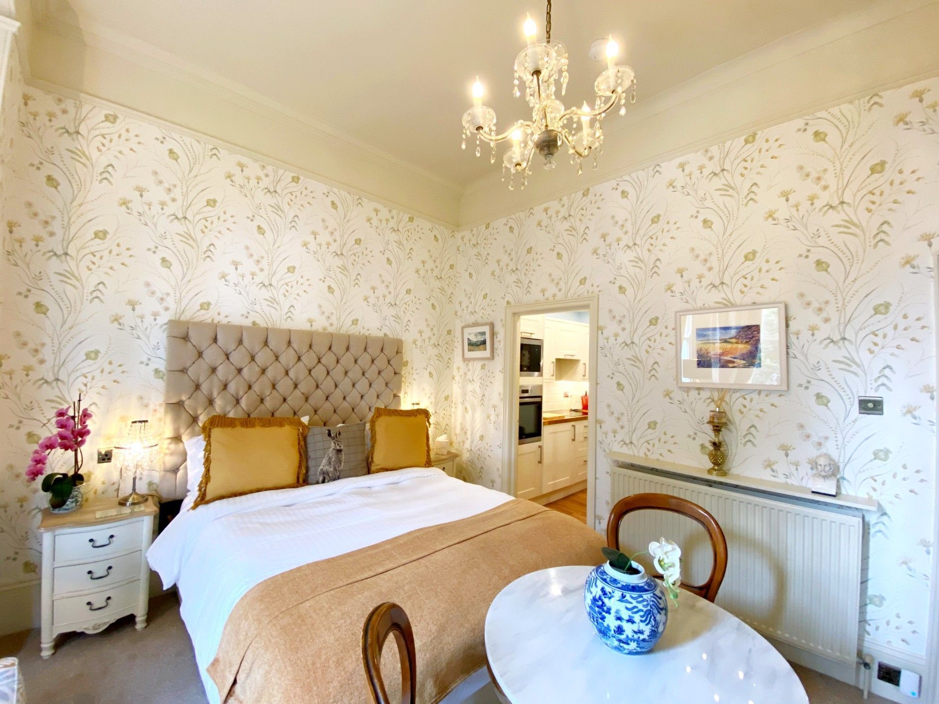 A bedroom with a tufted headboard, a bed with a white and brown bedspread, a bedside table with a vase, and a chandelier.