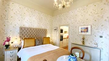 A bedroom with a tufted headboard, a bed with a white and brown bedspread, a bedside table with a vase, and a chandelier.