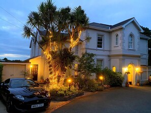 Een huis met twee verdiepingen, een oprit, een geparkeerde auto en een boom versierd met lichtjes.