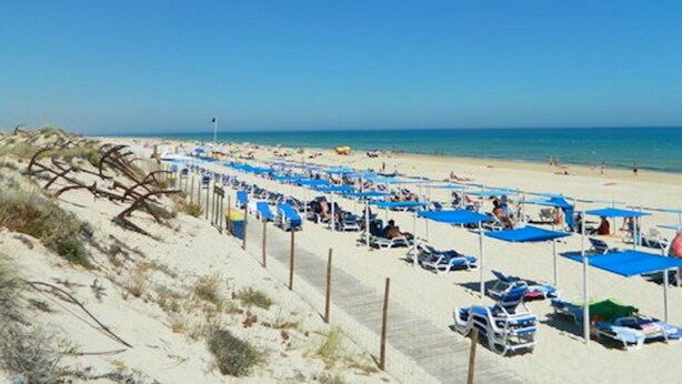 A beach with rows of blue umbrellas and sun loungers, sandy dunes, and the ocean in the background. - The White House - Tavira Historic old town center (Tavira)