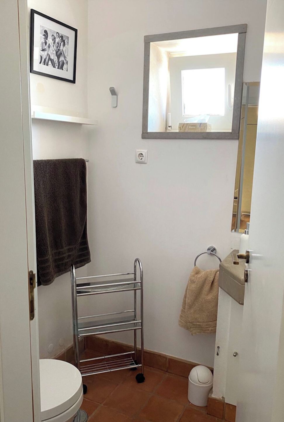 A compact bathroom with a toilet, a towel rack, a mirror, and a shelf.