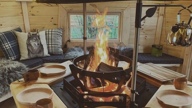 Un acogedor interior de cabaña con un estufa de leña en el centro, mesas de madera con cuencos y tazas, un sofá con cojines escoceses y una ventana con vista a follaje.