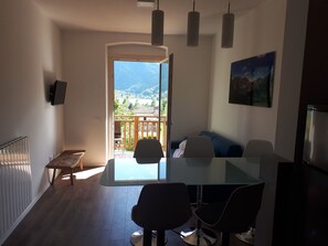 Un salon moderne avec une table à manger en verre, deux chaises, un canapé et une vue sur un balcon doté d’une terrasse en bois et donnant sur les montagnes.