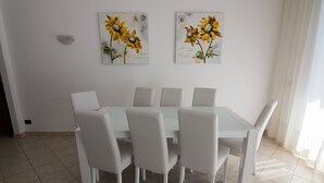 Un coin repas avec une table blanche et six chaises, deux tableaux représentant des tournesols accrochés au mur, et un interrupteur sur le mur.