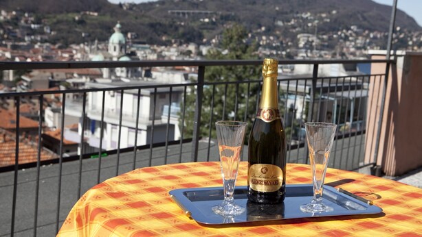 Une table de balcon avec une bouteille de vin Moscato et deux verres, donnant sur un paysage urbain avec des bâtiments et une montagne en arrière-plan.