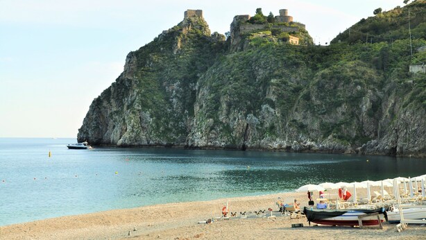 Una playa con barcos, un acantilado rocoso y una construcción que parece un castillo.