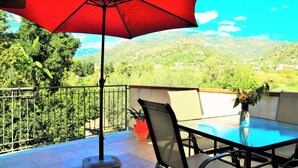 Un balcón con sombrilla roja, una mesa y sillas, con vistas a un paisaje montañoso.