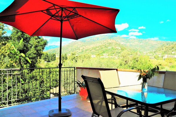 A balcony with a red umbrella, a table, and chairs, overlooking a mountainous landscape.
