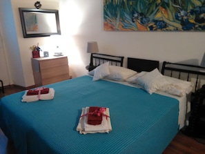 Uma cama bem arrumada com roupa de cama azul e almofadas brancas, uma mesa de cabeceira com abajur, uma cômoda e um quadro na parede.
