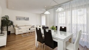 Un salon moderne avec un canapé blanc, une table à manger en verre et des chaises noires, une plante en pot et une œuvre d’art représentant un paysage urbain accrochée au mur.