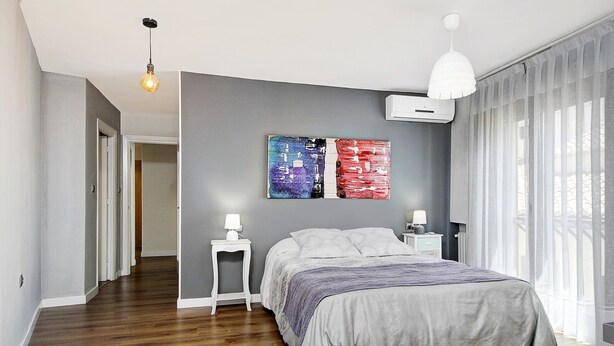 Une chambre moderne avec un grand lit, deux tables de chevet avec des lampes, un luminaire au plafond et une grande fenêtre ornée de rideaux légers.