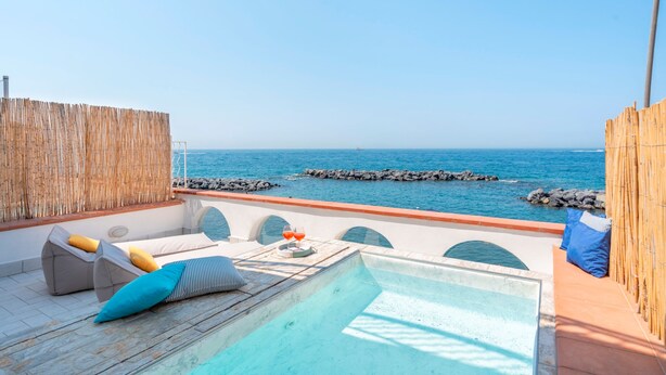 Zona de piscina con vista al mar, una valla de bambú y sillones de descanso.