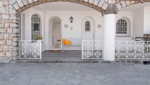 Un arco de piedra que conduce a una habitación blanca con una cama, un ventanal con celosía metálica y un aplique de pared.