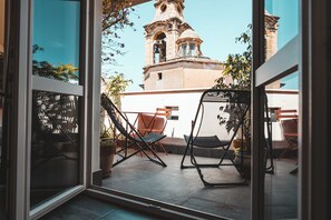 Vista a través de un ventanal a un patio amueblado al aire libre, con dos sillas y una hamaca, y un edificio con un campanario al fondo.