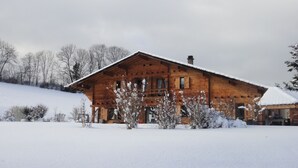 Una cabaña de madera con techo nevado, rodeada de terreno y árboles cubiertos de nieve.