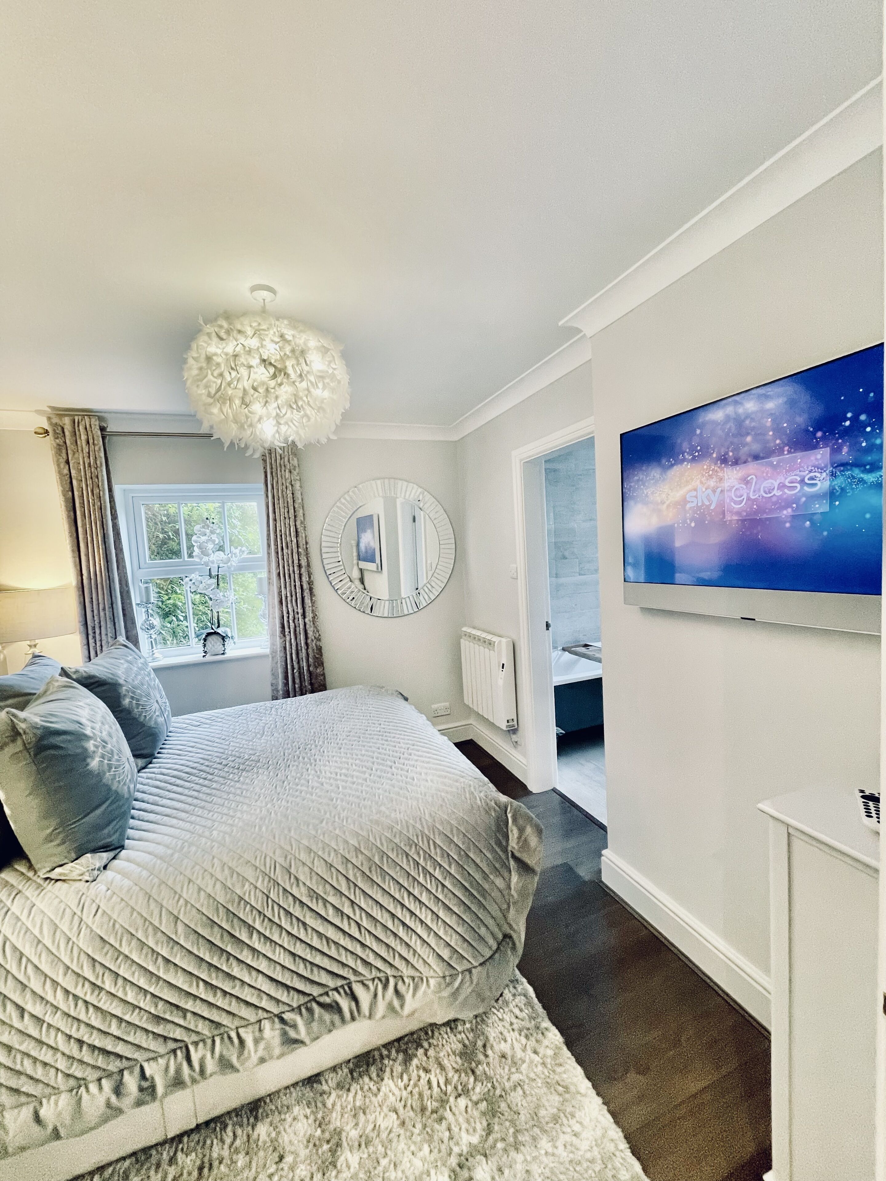 Quarto com cama, espelho redondo, lustre, janela e televisão fixada na parede.