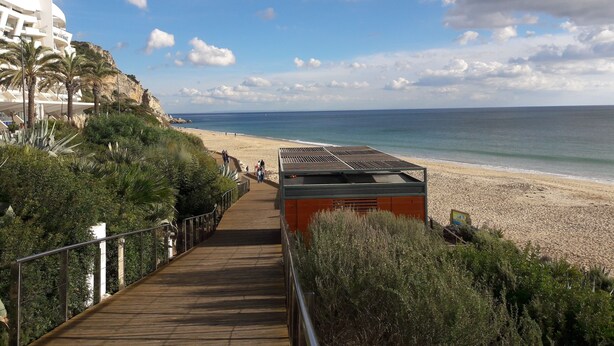 A coastal walkway leading to a beach with a rocky cliff and a modern building on the left.