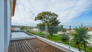 A balcony with a wooden floor, a glass railing, and a view of a green landscape with trees and buildings.