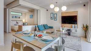 A modern living room with a dining area, a sofa set, a TV mounted on the wall, and a small side table with a potted plant. - Angels Suites (Platanias)