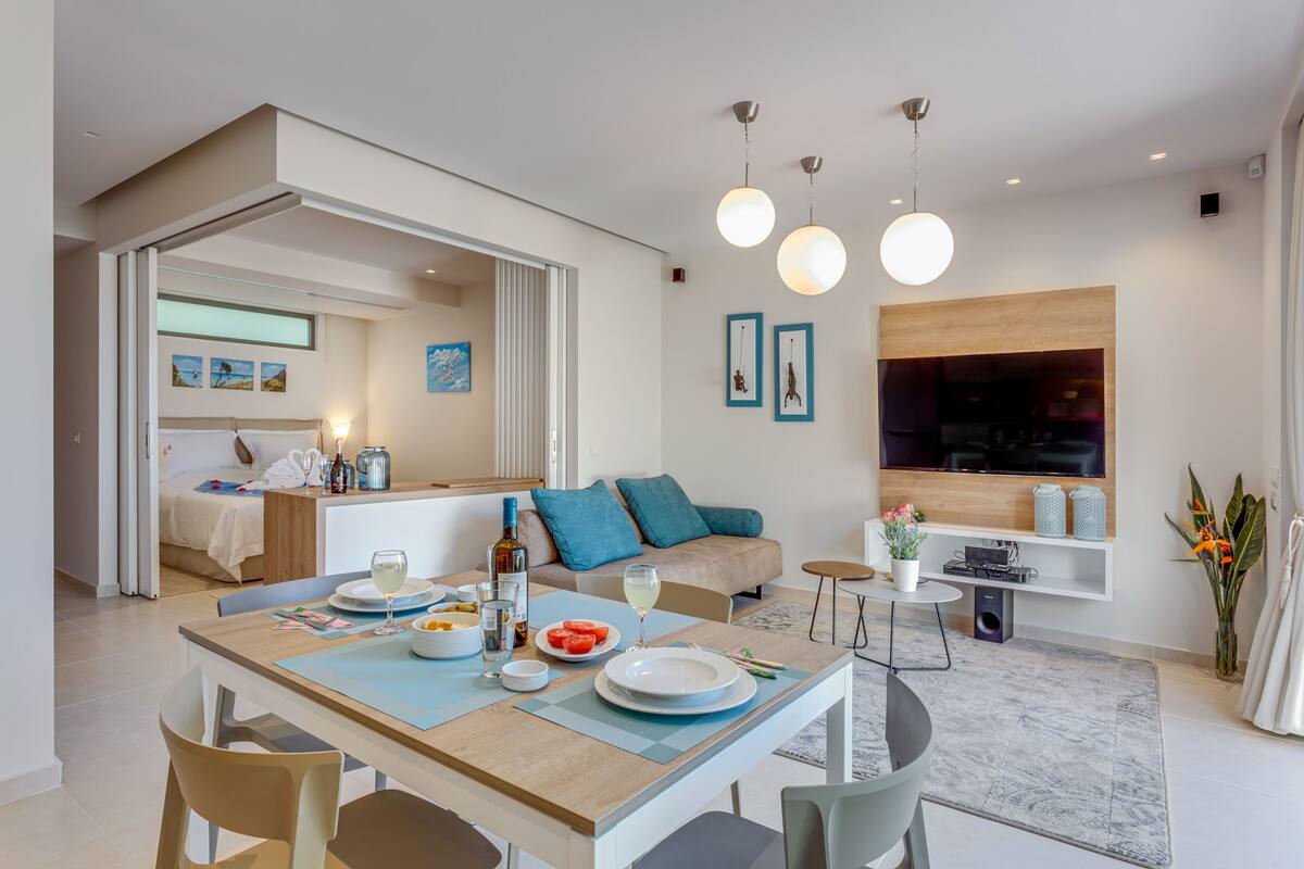 Un salon moderne comprenant un coin repas, un ensemble de canapés, un téléviseur fixé au mur et une petite table d’appoint agrémentée d’une plante en pot.