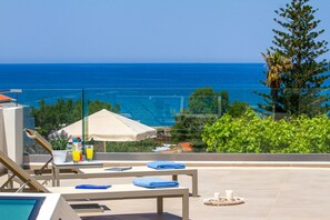 Zona de piscina con sillas de descanso, una mesa y toallas, con vista al océano.