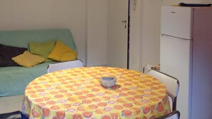 A small, well-lit room with a round table covered in a fruit-patterned tablecloth, a sofa with cushions, a white door, and a white refrigerator.